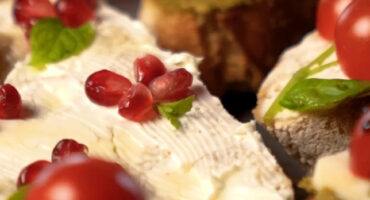 Close-up of bread slices spread with creamy cheese, topped with cherry tomatoes, pomegranate seeds, fresh mint, and a light honey drizzle