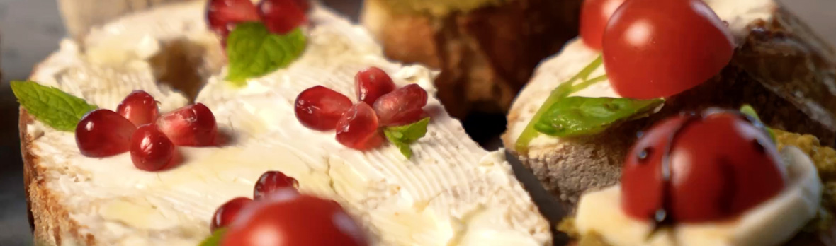 Close-up of bread slices spread with creamy cheese, topped with cherry tomatoes, pomegranate seeds, fresh mint, and a light honey drizzle