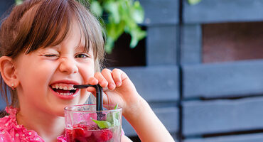 Adorable little girl gives a wink, drinking lemonade with raspberry and basil at table in cosy outdoor cafe