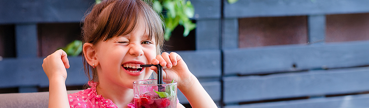 Adorable little girl gives a wink, drinking lemonade with raspberry and basil at table in cosy outdoor cafe