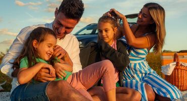 The father plays with his daughter while the mother fixes her other daughter's hair, the family spends quality time outside