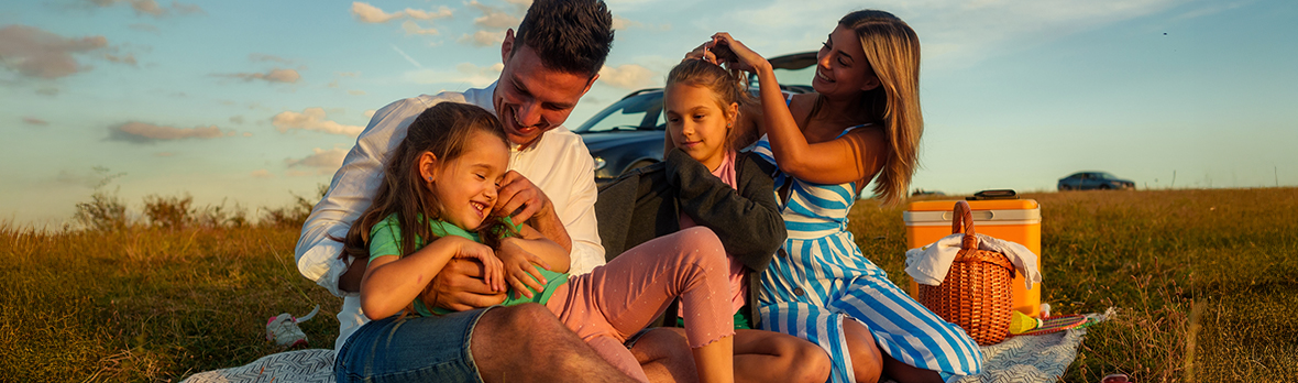 The father plays with his daughter while the mother fixes her other daughter's hair, the family spends quality time outside
